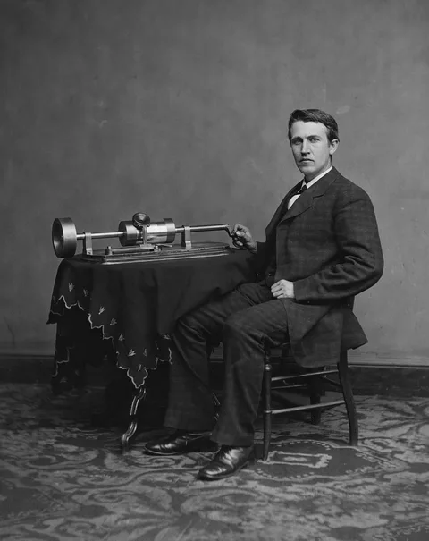 Thomas Edison seated beside a tinfoil cylinder phonograph, with his hand on its crank.