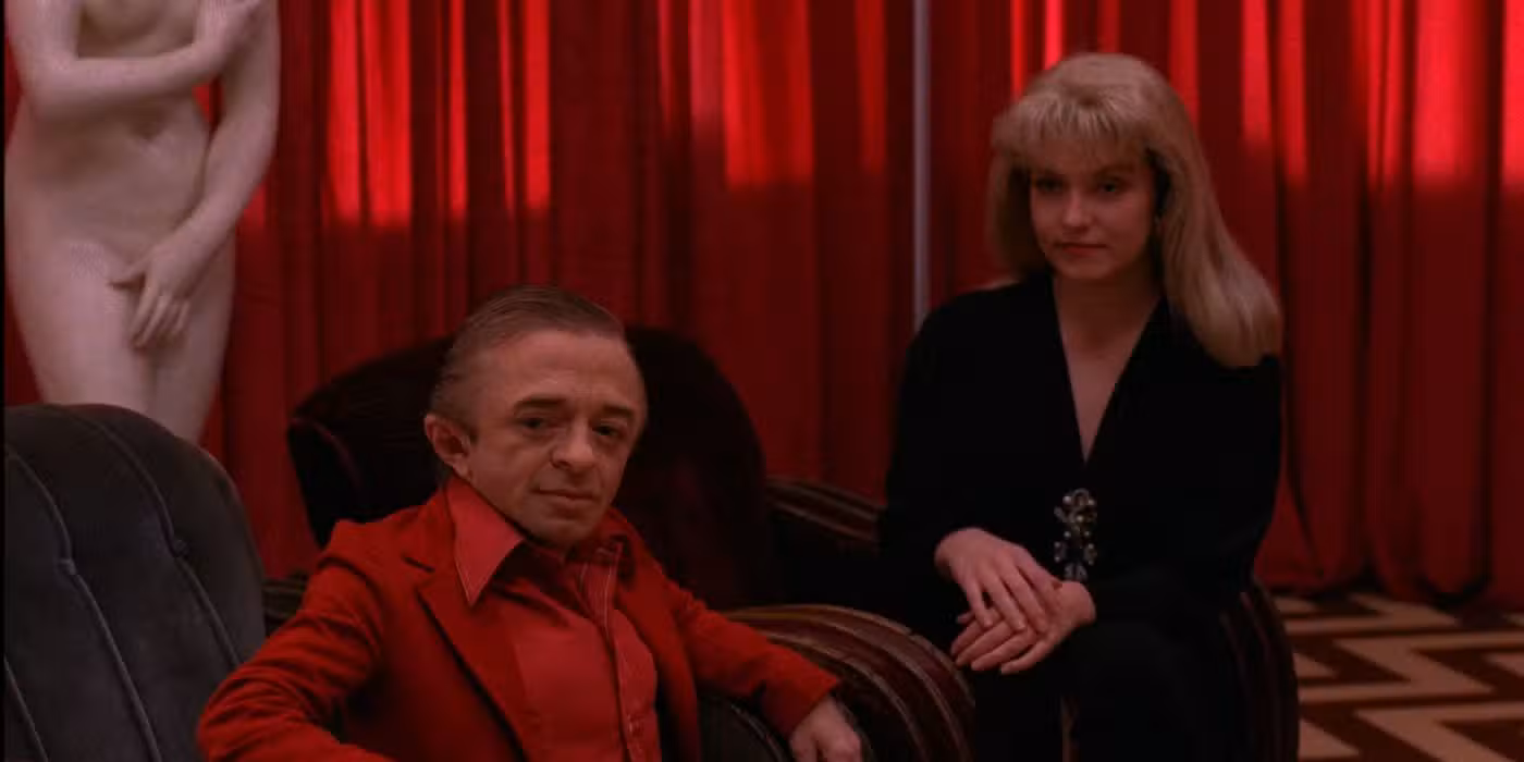 A man in a red suit and a woman in black seated in the Red Room, with red curtains and a statue behind them.